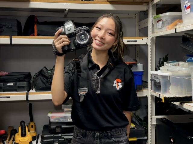 Pham in black polo holding DSLR in an equipment storage room