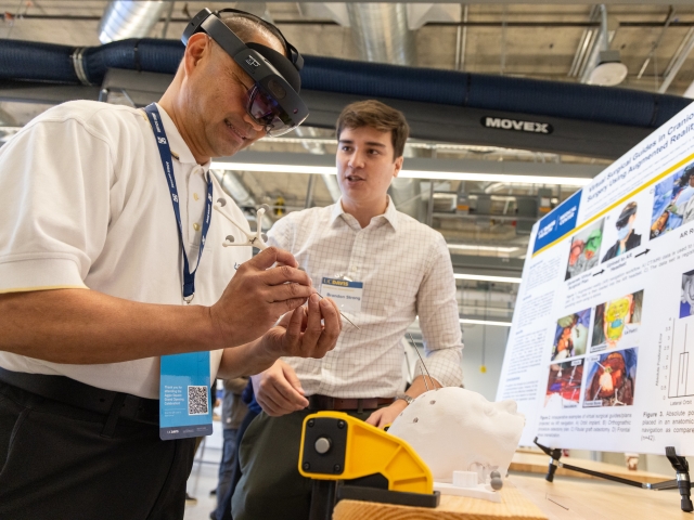 One person wears augmented reality goggles while another stands close by near a poster and interactive activity