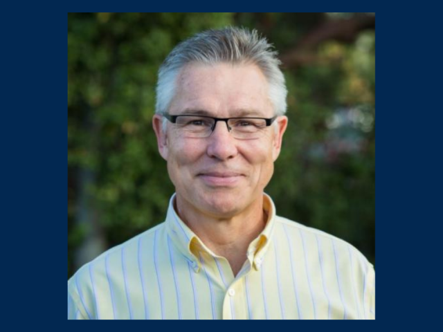 A headshot of Simon Bare, a person with short gray hair and glasses, wearing a light yellow button-up shirt with thin blue stripes. He is smiling gently, and the background features blurred greenery. The image is framed with a dark blue border.