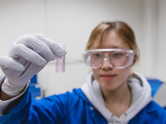Young scientist in lab wearing goggles and gloves holding small vial with pink liquid — photograph