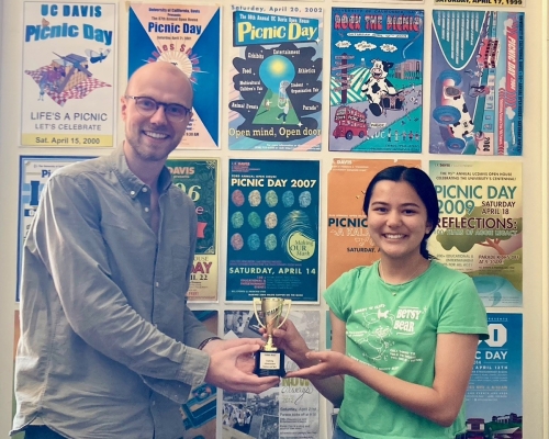 Two UC Davis QCAD members pose for a photo in front of Picnic Day posters, holding a trophy