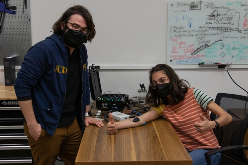 One person stands and one person sits at table with arms hooked up to machine