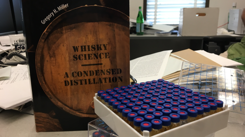 Book with title Whisky Science a Condensed Distillation" sits on desk near vials of liquid chemical.