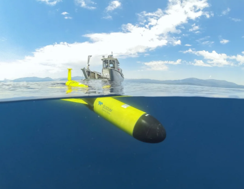 Bright yellow autonomous underwater vehicle half in blue water, just launched from a boat in the background of the image