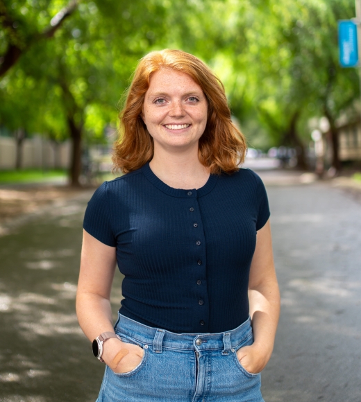 Headshot photograph of Christina Harvey, hands in pockets on tree-lined street