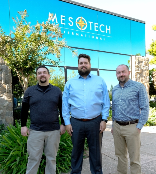 Three people wearing business casual attire in front of an office building with the Mesotech International, Inc. logo on blue glass.