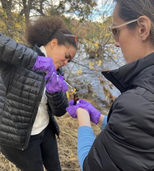 Jasquelin Peña wears gloves and handling a sample outdoors, with another person standing close holding a sample container