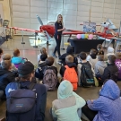 Students sitting on the ground while paying attention to a speaker near an airplane in a museum