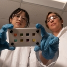 Two researchers stand in a lab looking at a photocell, wearing PPE