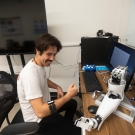 Person sits at a desk wearing a mechanical arm band and looking at a prosthetic arm