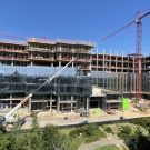 A large construction site featuring a multi-story building in progress under a clear blue sky. The structure has a glass facade on the lower levels and exposed steel framing on the upper levels. Two tall red cranes tower over the site, and construction equipment, including a crane and other vehicles, is visible on the ground. Surrounding greenery frames the scene, with additional buildings and trees in the background.