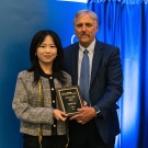 Peifen Lyu stands to the left of Dean Richard Corsi, holding an award in front of a blue backdrop