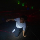 A person crawls on the floor, surrounded by colorful balloons and red laser beams in a dim setting.