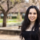 A woman with long dark hair wearing a dark top stands in front of a natural background