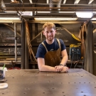 A young man with red hair wears an apron and stands in a welding studio