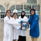 Four students in lab coats hold a cube structure in front of a building with windows