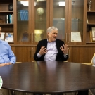 Three individuals are seated at a round wooden table engaged in a discussion. Behind them, a wooden cabinet displays books, awards, and a framed photograph. The person in the center gestures with open hands, emphasizing a point, while the others listen attentively. In the lower-left corner, a circular logo with a globe design and the text 'Engineering Progress 2024-2025' is visible.