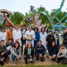 A large group of Chem-E-Car members stand together outdoors near a sculpture, one person in the middle holds the car