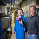 Two researchers hold a pink device standing in front of large tanks in a laboratory