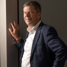 Simon Cherry wears a suit and stands with one hand against the wall