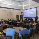 An audience listens attentively as Dean Corsi stands at a podium with a UC Davis banner, speaking at the 'Tensions at the Edge' symposium. The event is held in a spacious conference room with round tables and a large projection screen displaying the symposium title.