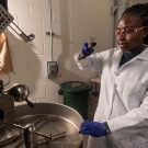 A black female researcher wearing a white lab coat and blue latex gloves removes a sample of coffee beans from a Probat coffee roaster