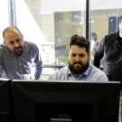 Three engineers looking at two computer monitors in an office with a large window. 