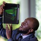alt="Ismael Mayanja wearing a suit holds up a tablet with a green model displayed"
